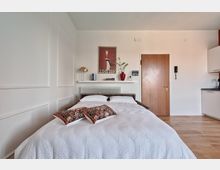 Camera da letto moderna e luminosa con letto matrimoniale, pareti bianche con boiserie e pavimento in legno. Dietro al letto ci sono una mensola con lampada e libri, un poster, un radiatore e una porta d’ingresso; a destra si vede un angolo cottura con pensili bianchi e piano di lavoro scuro.