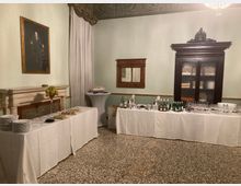 Sala interna in stile classico con pavimento in graniglia, pareti chiare e soffitto decorato, con camino in marmo e ritratto appeso. Lungo le pareti sono allestiti tavoli da buffet con tovaglie bianche, bicchieri e bottiglie, accanto a un grande armadio in legno e una finestra con tende.