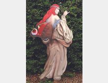 Statua dipinta in un giardino, raffigurante una figura femminile in abito lungo con capelli rossi e grandi ali di farfalla. Sullo sfondo si vede una fitta siepe verde e il terreno è coperto di ghiaia.