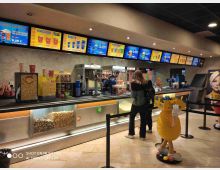 Banco bar di un cinema per snack, con menu digitali illuminati sopra il bancone e macchine per popcorn, nachos e bibite sul piano di lavoro. Davanti ci sono corde segnapercorso e un’esposizione di caramelle e contenitori trasparenti pieni di popcorn e dolci.