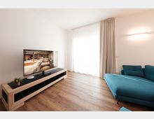 Soggiorno moderno e minimalista con pavimento in legno, pareti bianche, mobile TV basso con televisore e grande finestra con tende chiare fino a terra. A destra si vede un divano angolare blu sotto una lampada a parete.