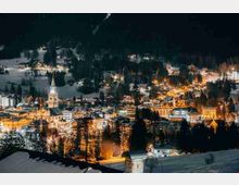 Veduta serale dall’alto di una cittadina alpina con case e palazzi illuminati, dominata da una chiesa con alta guglia centrale. Intorno si estendono pendii innevati e boschi scuri che circondano l’abitato.