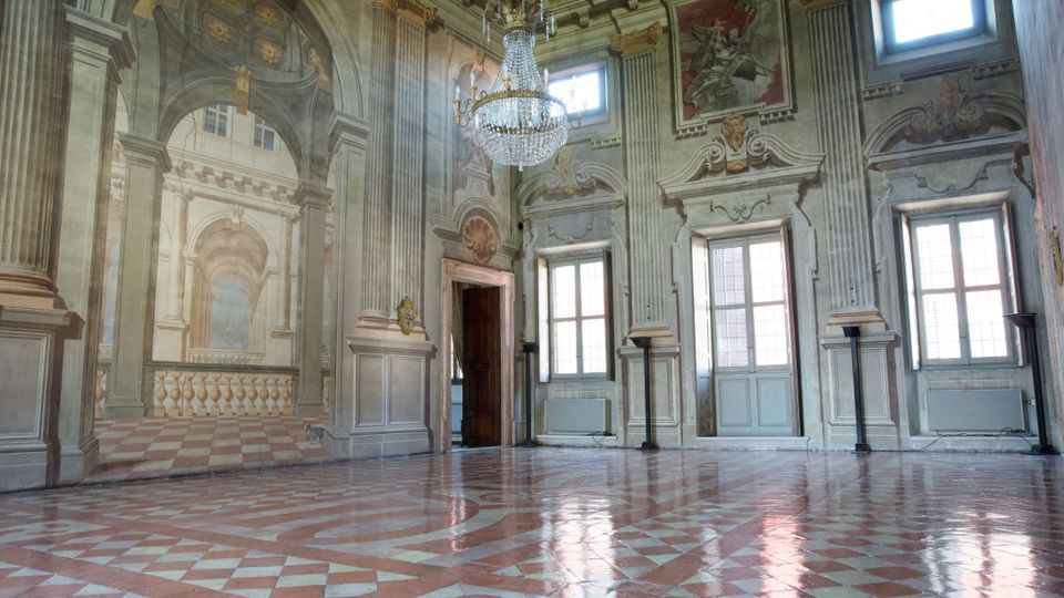 Interno di una sala storica con un soffitto decorato, pareti affrescate con motivi architettonici illusionistici, alti pilastri e ampie finestre. Al centro è sospeso un grande lampadario di cristallo, mentre il pavimento ha un motivo geometrico in marmo rosso e bianco.