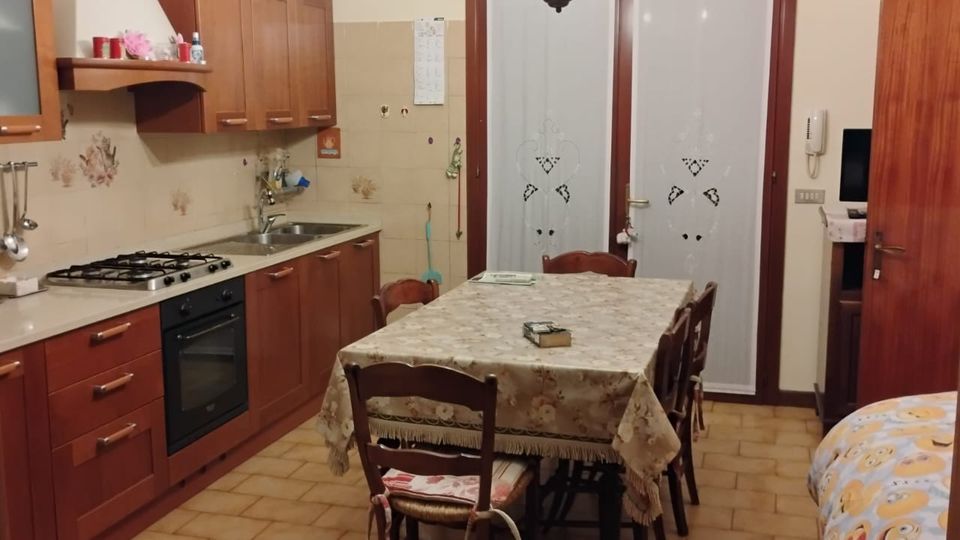 Cucina abitabile con mobili in legno, piano cottura e forno incassato lungo la parete sinistra, lavello e pensili. Al centro c’è un tavolo rettangolare con tovaglia e sedie, con pavimento in piastrelle e porte a vetro sul fondo.