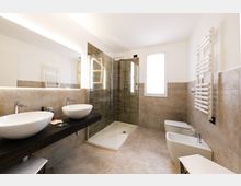 Bagno moderno con piastrelle beige e doccia angolare in vetro, doppio lavabo su piano scuro con grande specchio illuminato. Presenti finestra, termoarredo a parete, WC e bidet su pavimento in gres.