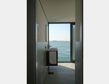 Interno di un bagno moderno e minimale con lavabo sospeso e rubinetteria nera, affacciato su una grande porta-finestra. Attraverso l’apertura si vede direttamente il mare calmo e l’orizzonte in lontananza.