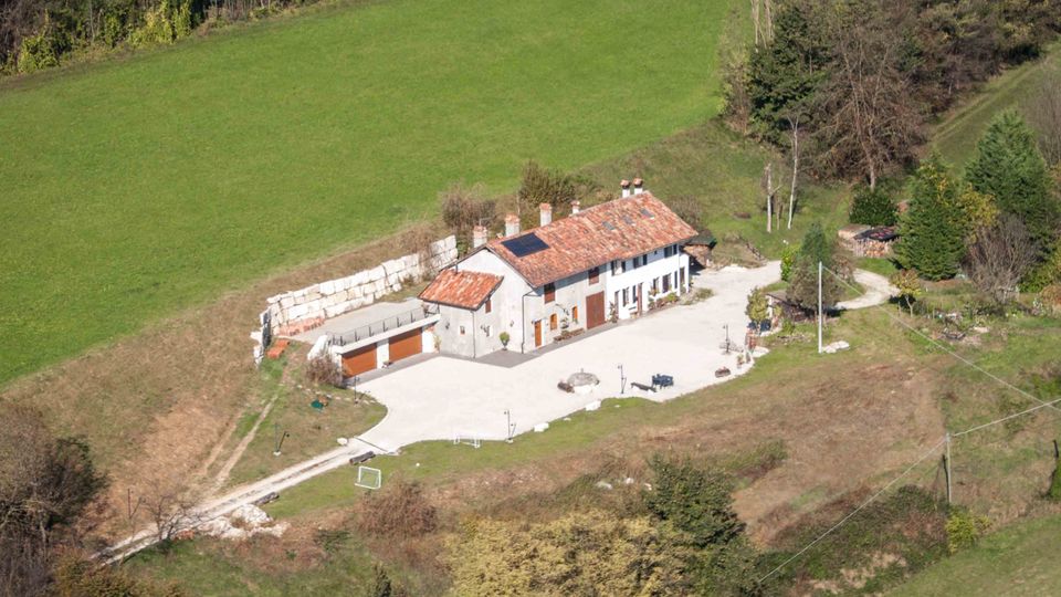Veduta aerea di una casa rurale isolata con tetto in tegole rosse e cortile pavimentato, affiancata da un annesso con autorimessa. L’edificio è circondato da prati verdi e bosco su un pendio collinare, con strada sterrata di accesso.