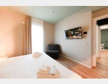 Camera da letto moderna con letto matrimoniale, pavimento in legno e pareti dai toni chiari; una grande finestra con tende lascia entrare luce naturale. Sulla parete è montata una TV, accanto a una poltrona scura e a una porta che conduce a un’altra stanza.