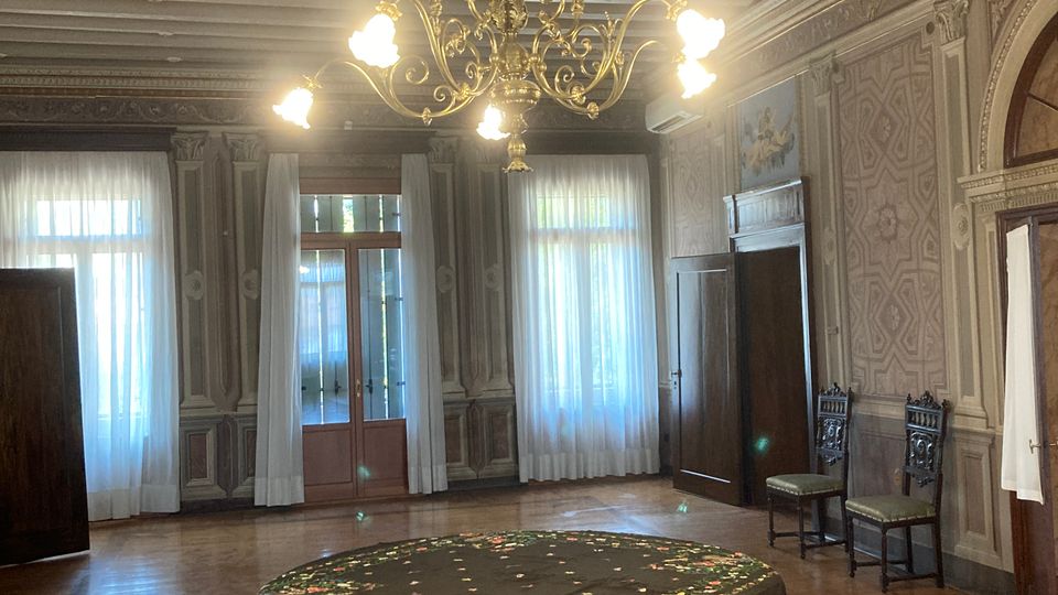Salone storico con pavimento in legno e soffitto a travi dipinte, illuminato da un grande lampadario in ottone. Al centro c’è un tavolo rotondo con tovaglia ricamata e frange, circondato da pareti ornate e alte finestre con tende bianche.
