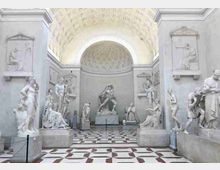 Interno di una sala museale neoclassica con pavimento a scacchi e volte a cassettoni, delimitata da pareti in pietra chiara. Lungo i lati sono esposte numerose sculture in marmo su piedistalli e in nicchie, con una statua centrale in fondo alla sala.