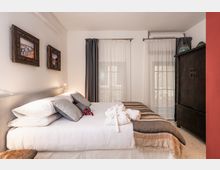 Camera da letto luminosa con grande letto matrimoniale, testiera imbottita e cuscini, con coperta a righe e asciugamani piegati sul letto. Sul fondo ci sono due alte finestre con tende bianche e grigie; a destra un grande armadio scuro e alle pareti quadri con cornici intagliate.