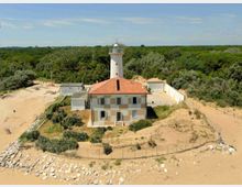 Veduta aerea di un faro con torre bianca e edificio annesso con tetto in tegole rosse, circondato da dune sabbiose e vegetazione costiera. Intorno si vedono recinzioni, piccoli edifici di servizio e una vasta area di bosco sullo sfondo.