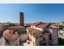 Vista dall’alto di un complesso religioso con tetti in coppi e un alto campanile in mattoni, affacciato su un cortile interno con portico ad archi. Sullo sfondo si estende il centro urbano con edifici bassi e tetti rossi sotto un cielo sereno.