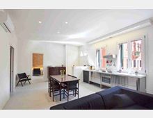 Interno di un appartamento moderno e luminoso con cucina a vista lineare lungo la parete destra, grandi finestre e pensili in metallo. Al centro c’è un tavolo da pranzo in legno scuro con sedie, mentre in primo piano si vede un divano e sul fondo un’area separata con parete in mattoni a vista.