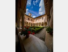Chiostro in mattoni con portici ad archi sorretti da colonne in pietra, affacciati su un giardino centrale con siepi geometriche e vialetti in ghiaia. Le pareti mostrano tracce di affreschi e finestre ad arco, con cielo azzurro visibile sopra il cortile.
