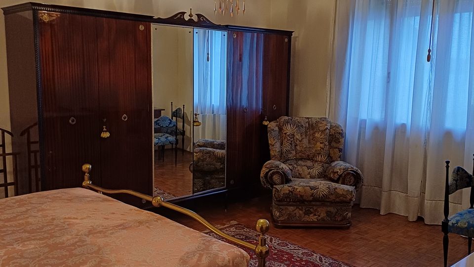 Camera da letto con arredi classici: letto con struttura in ottone, grande armadio in legno scuro con ante a specchio e lampadario a gocce sospeso. Accanto a una finestra con tende leggere c’è una poltrona imbottita, con tappeti sovrapposti su pavimento in parquet.