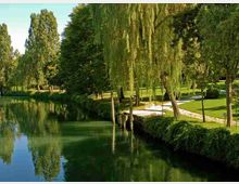 Parco alberato lungo un canale o fiume calmo, con acqua verde e riflessi degli alberi. Sulla riva si vedono salici piangenti, prato curato, siepi basse e un sentiero con piccole piattaforme in legno vicino all’acqua.
