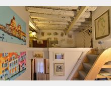 Interno di una mansarda con soppalco in legno e soffitto a travi bianche, raggiunto da una scala curva rivestita in moquette. Le pareti sono decorate con numerosi quadri e stampe, e il soppalco ospita mobili e oggetti d’arredo con una grata in legno sul fondo.