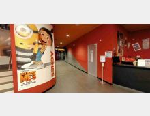 Ingresso di un cinema con corridoio lungo e pareti rosso scuro, pavimento grigio e illuminazione a soffitto. A sinistra c’è una grande locandina pubblicitaria su una colonna curva, mentre a destra si vede un bancone con monitor e poster alle pareti.