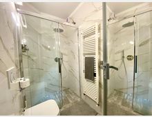 Bagno moderno con due box doccia in vetro e profili metallici, rivestimenti effetto marmo bianco e pavimento grigio. Al centro c’è uno scaldasalviette verticale bianco; in primo piano si vede parte del WC.
