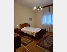 Camera da letto con letto matrimoniale in legno e copriletto chiaro, comodino con lampada e due tappeti decorati su pavimento in parquet. Una grande finestra con tende bianche illumina la stanza, con lampadario a soffitto e pareti chiare.
