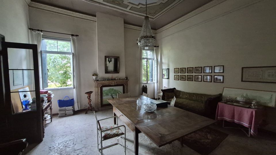 Interno di un salone d’epoca con soffitto alto decorato a stucchi e lampadario in cristallo, pavimento in marmo e grandi finestre con tende bianche. Al centro c’è un tavolo in legno, lungo una parete un divano e una fila di cornici, e in fondo un camino con specchio.