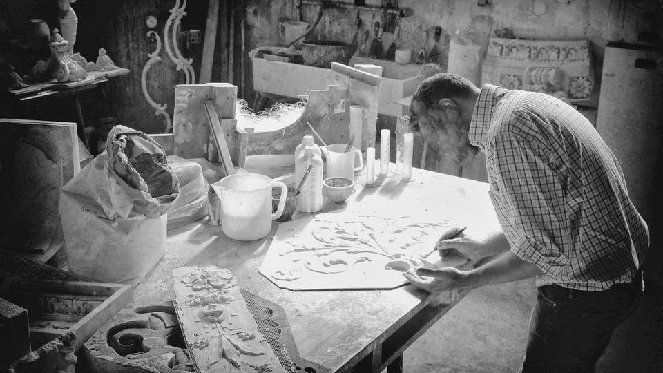 Interno di un laboratorio artigianale con grande banco da lavoro coperto di polvere, stampi decorativi e utensili, tra contenitori e flaconi. Sullo sfondo si vedono lavandino, mensole con attrezzi e pannelli ornamentali appoggiati alle pareti.