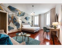 Camera da letto moderna con pavimento in legno lucido, grande letto matrimoniale e parete decorata con un murale di foglie. Due ampie finestre con tende chiare illuminano una zona scrivania con sedia e TV a parete, con tavolini bassi su tappeto verde in primo piano.