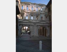 Facciata di un edificio storico in pietra con due grandi archi di passaggio e ordini di finestre ad arco sovrapposte, con fregi e frontoni classici. Inquadratura su una strada urbana acciottolata che attraversa gli archi, tra edifici ai lati.