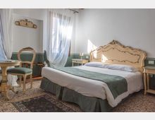 Camera da letto luminosa con letto matrimoniale e testiera intagliata, pareti bianche con fascia verde e pavimento in graniglia con tappeto. Una finestra con tende leggere illumina comodini e piccolo tavolo con sedia in stile classico.