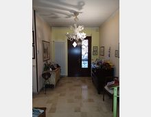 Ingresso di abitazione con pavimento in pietra chiara, lampadario in vetro al soffitto e porta d’ingresso in legno scuro con inserto in vetro decorato. Lungo le pareti ci sono un radiatore verticale, mobili bassi con cornici e oggetti decorativi e quadri appesi.