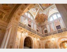 Interno di un palazzo storico con loggiato e balaustre in pietra, grandi arcate e pareti e soffitti decorati da affreschi. In alto si vedono finestre ad arco che illuminano lo spazio e lampadari sospesi.