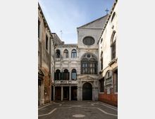 Piccolo cortile lastricato tra edifici storici con facciate in intonaco e mattoni a vista. Sul fondo si vede una facciata in stile veneziano con archi, portale e grande finestra con inferriate, sormontata da un frontone con croce e oculo circolare.