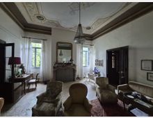 Salone di una casa d’epoca con soffitto decorato a stucchi e lampadario a gocce, camino in marmo con grande specchio e ampie finestre con tende bianche. Arredi classici con poltrone e divani, tavolini, scrivania e porte scure su pavimento chiaro.