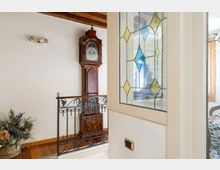 Disimpegno interno con pavimento in legno, ringhiera in ferro battuto e soffitto con travi a vista. Sulla parete è presente un grande orologio a pendolo in legno e una finestra con vetro decorato a rombi colorati.