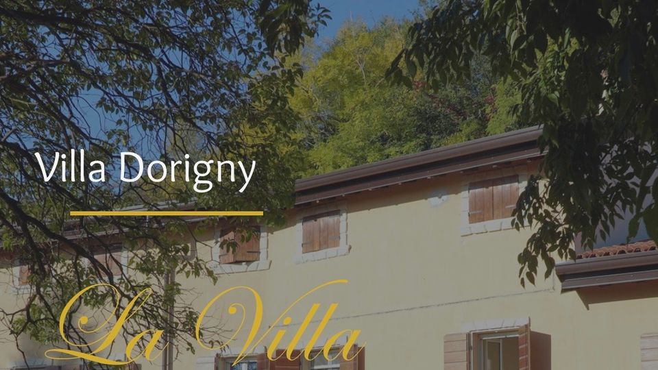 Facciata di una villa di campagna a due piani con intonaco chiaro e persiane in legno, con finestre rettangolari e tetto a falde. L’edificio è parzialmente incorniciato da rami e alberi, con vegetazione sullo sfondo.