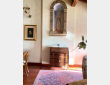 Interno di una stanza in stile classico con pareti chiare, pavimento in legno e tappeto decorato; al centro un mobile scrittoio in legno sormontato da una nicchia ad arco scolpita con affresco. Sulle pareti sono presenti un quadro incorniciato e un’applique, con travi a vista in alto e arredi parzialmente visibili ai lati.