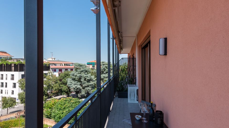 Balcone lungo e stretto di un appartamento con pavimento piastrellato, ringhiera metallica scura e parete esterna color pesca con porta finestra. In fondo si vede un piccolo tavolo con sedie e, oltre la ringhiera, una vista su alberi e palazzi sotto un cielo sereno.