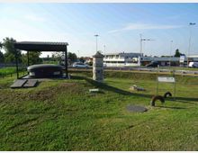 Area verde lungo una strada trafficata con un piccolo padiglione dal tetto metallico che copre una struttura circolare bassa e scura, accanto a un pilastro in pietra e a pannelli informativi. Sullo sfondo si vedono edifici commerciali, lampioni e una rotatoria con auto in transito.