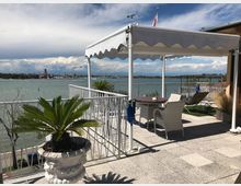 Terrazza sul lungolago con pavimentazione in pietra, ringhiera bianca e pergola con tenda, arredata con tavolo e sedie e piante in vaso. Sullo sfondo si vede lo specchio d’acqua con barche e una città costiera all’orizzonte sotto un cielo parzialmente nuvoloso.