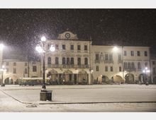 Piazza cittadina di notte coperta di neve, con un edificio storico a più piani sullo sfondo dotato di portici ad archi, balconi e un orologio centrale in facciata. Lampioni accesi illuminano la scena mentre la neve cade fitta sul pavimento innevato.