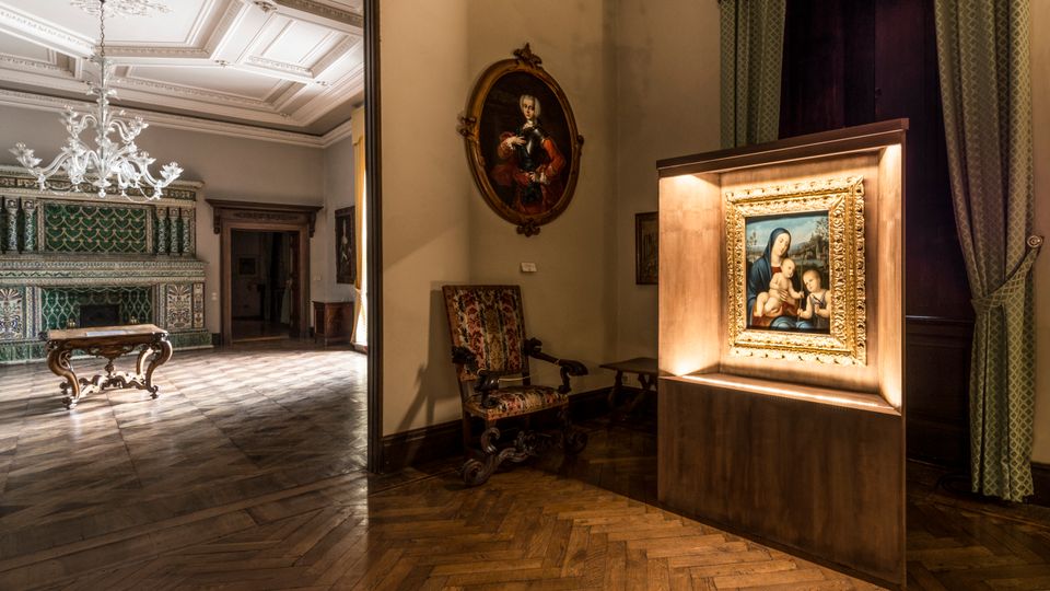 Interno di una sala museale in un palazzo storico, con pavimento in parquet a spina di pesce, soffitti decorati e lampadari in vetro. In primo piano una teca illuminata con un dipinto incorniciato, accanto a una poltrona antica, mentre sullo sfondo si vede un grande camino in maiolica verde e un tavolo intagliato.