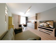 Soggiorno open space moderno con cucina lineare a parete, tavolo rotondo con sedie vicino a una grande finestra con tende e divano in primo piano. A destra ci sono un mobile TV basso con televisore, mentre a sinistra un armadio in stile rustico; pavimento in legno chiaro e illuminazione a binario sul soffitto.