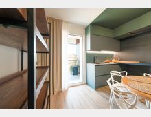 Cucina moderna con mobili verde salvia e grigi, piano di lavoro con lavello e illuminazione sottopensile, affiancata da un tavolo rotondo in legno con sedie bianche. Una portafinestra con tende chiare si apre su un balcone con vaso e ringhiera, mentre a sinistra si vede una libreria in legno e metallo.