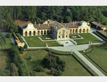 Veduta aerea di una villa storica con corpo centrale e ali laterali ad arcate, tetti in coppi e facciate simmetriche. Davanti si trovano un giardino geometrico con aiuole e un ampio viale d’accesso, circondati da prati e bosco sullo sfondo.