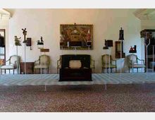 Sala espositiva tipo museo con pareti bianche e pavimento in graniglia, con sedie d’epoca allineate lungo le pareti e vari oggetti e dispositivi storici esposti su piedistalli. Al centro si trova una vetrina scura e, sulla parete di fondo, un grande quadro decorativo sopra la disposizione degli oggetti.