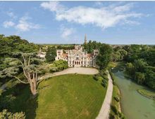 Veduta aerea di una villa storica in stile neogotico con facciata simmetrica e tetti in laterizio, circondata da un ampio parco alberato. In primo piano si vedono un grande prato curato, un viale sterrato e un corso d’acqua che costeggia il giardino.