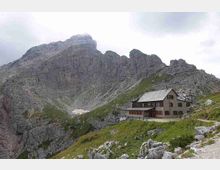 Rifugio alpino in muratura con tetto a falde e persiane in legno, situato su un pendio erboso e roccioso. Sullo sfondo si alzano alte pareti montuose grigie e frastagliate sotto un cielo nuvoloso.