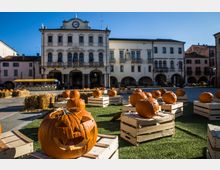 Piazza cittadina con un palazzo storico a più piani con torretta e orologio sulla facciata, affiancato da edifici con portici ad arcate. In primo piano, cassette di legno e balle di fieno disposte su un’area verde con numerose zucche intagliate.