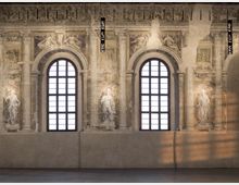 Interno di una sala storica con due alte finestre ad arco e griglie scure, incorniciate da colonne e decorazioni murali affrescate con figure e motivi classici. Sulla parete superiore sono visibili elementi ornamentali e due corpi illuminanti sospesi.